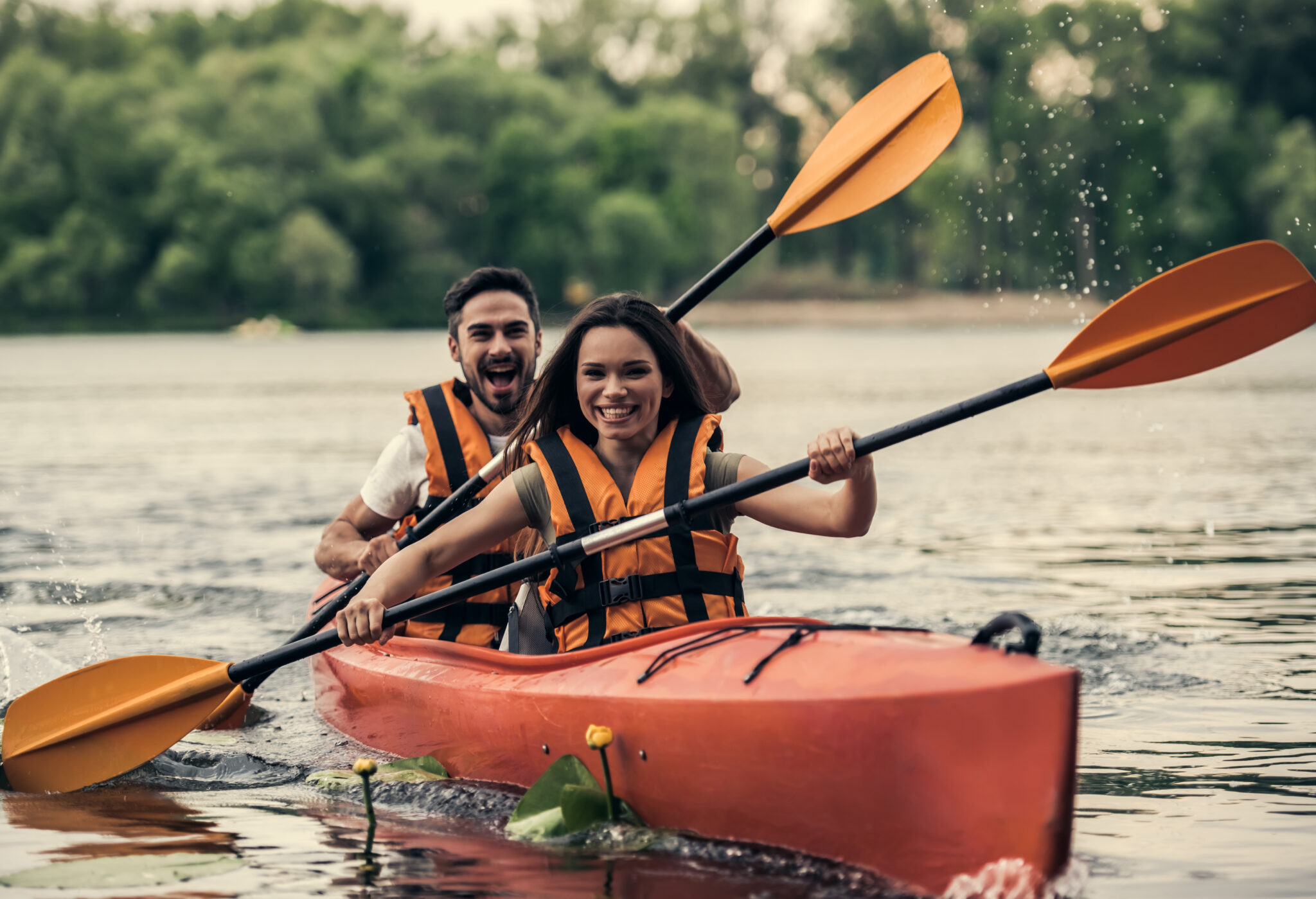 A joyful couple kayaks on a lake in orange life vests. They paddle energetically, splashing water, surrounded by calm water and green trees.