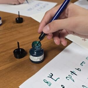 Ръка потапя калиграфско перо в синя мастилница върху дървена маса по време на уъркшоп.